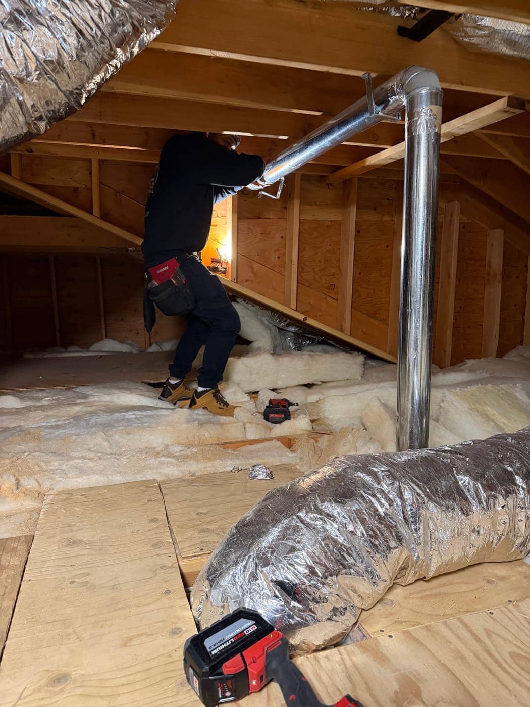 A technician in an unfinished attic fastening a rigid metal vent pipe, with silver flexible ducts lying on the floor boards.