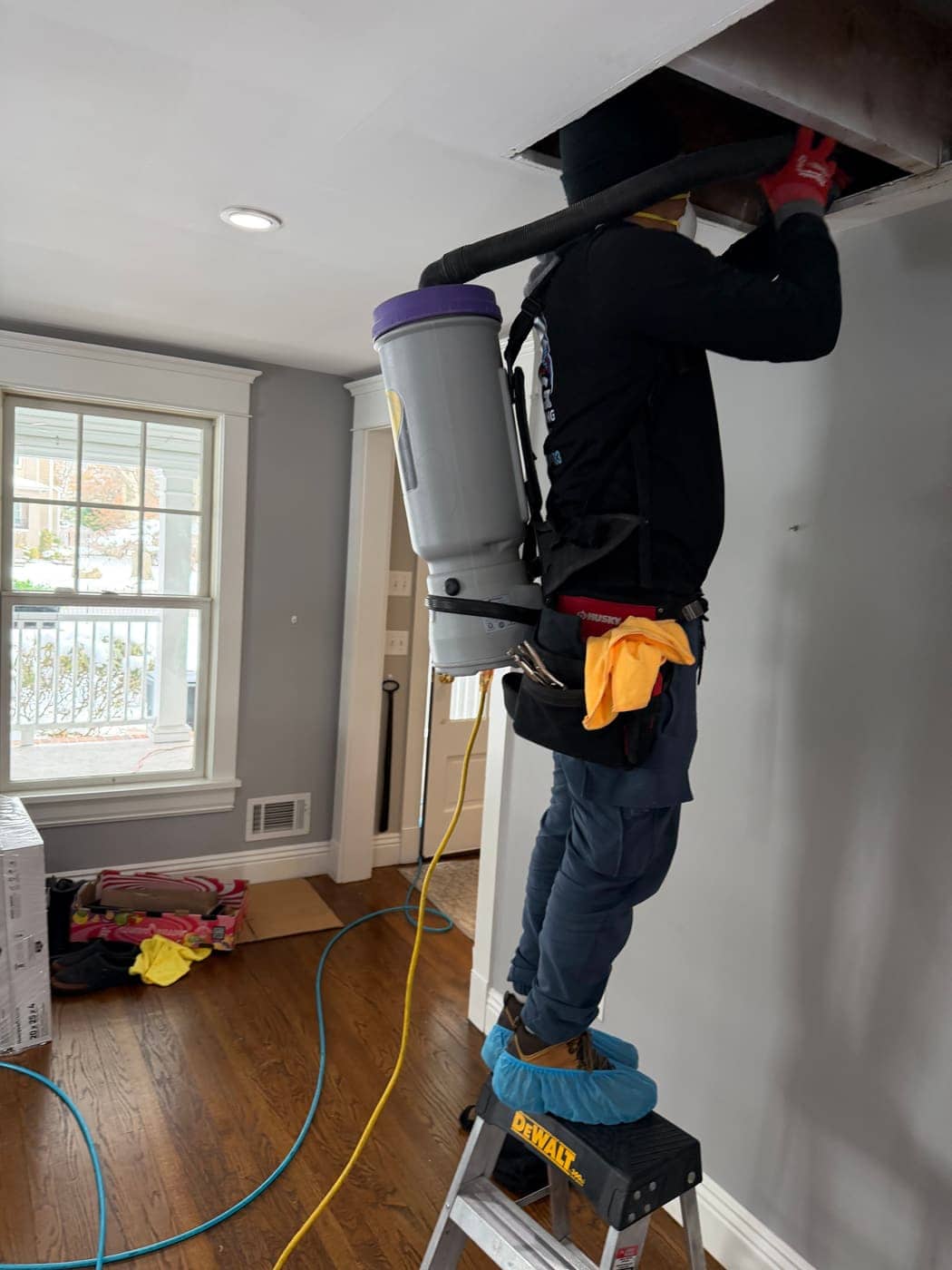 A technician on a step stool, wearing a backpack vacuum, working on an opening in a ceiling while plastic covers protect his shoes.