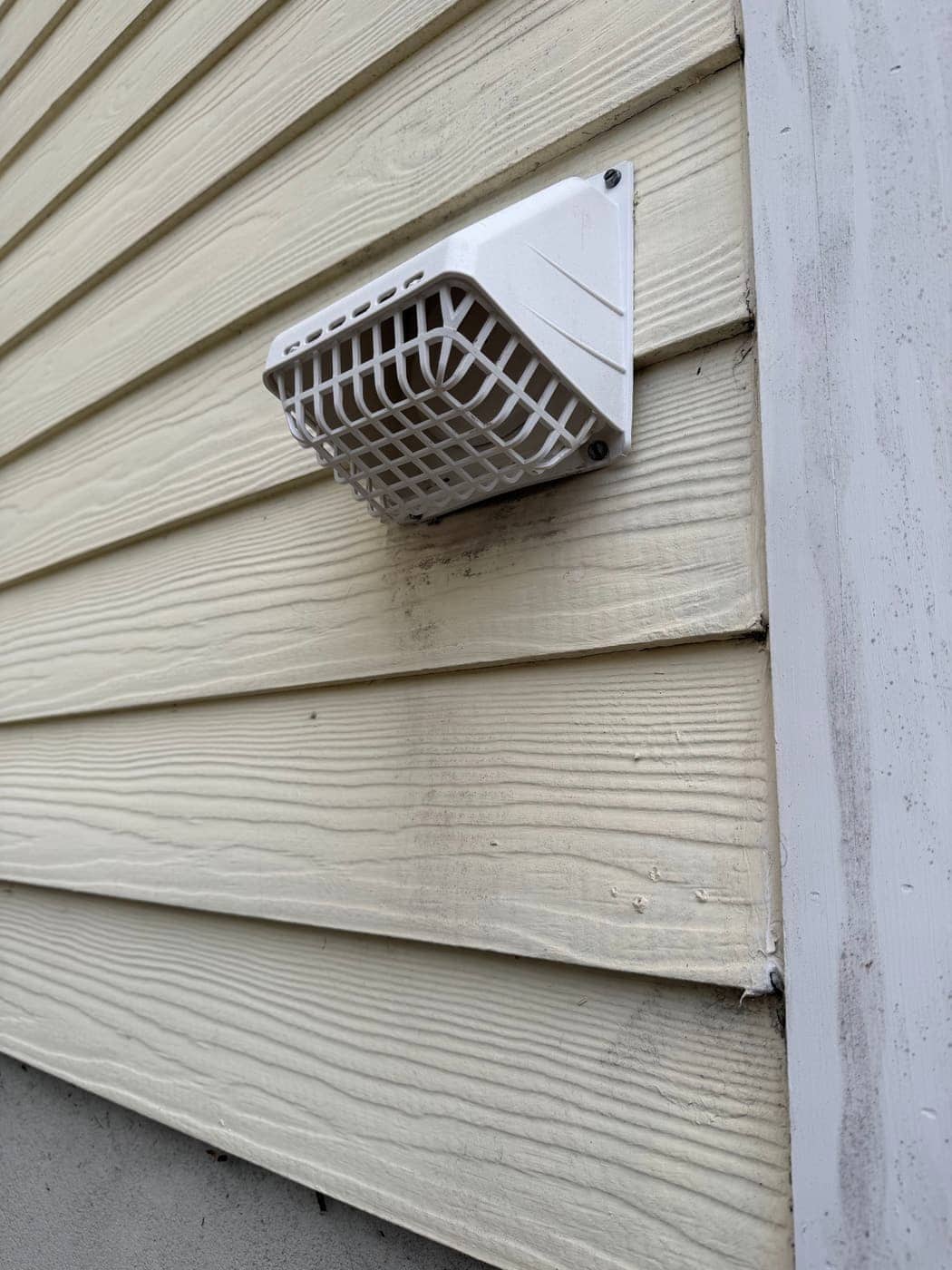 A white dryer vent hood with a guard grille, mounted on beige siding.