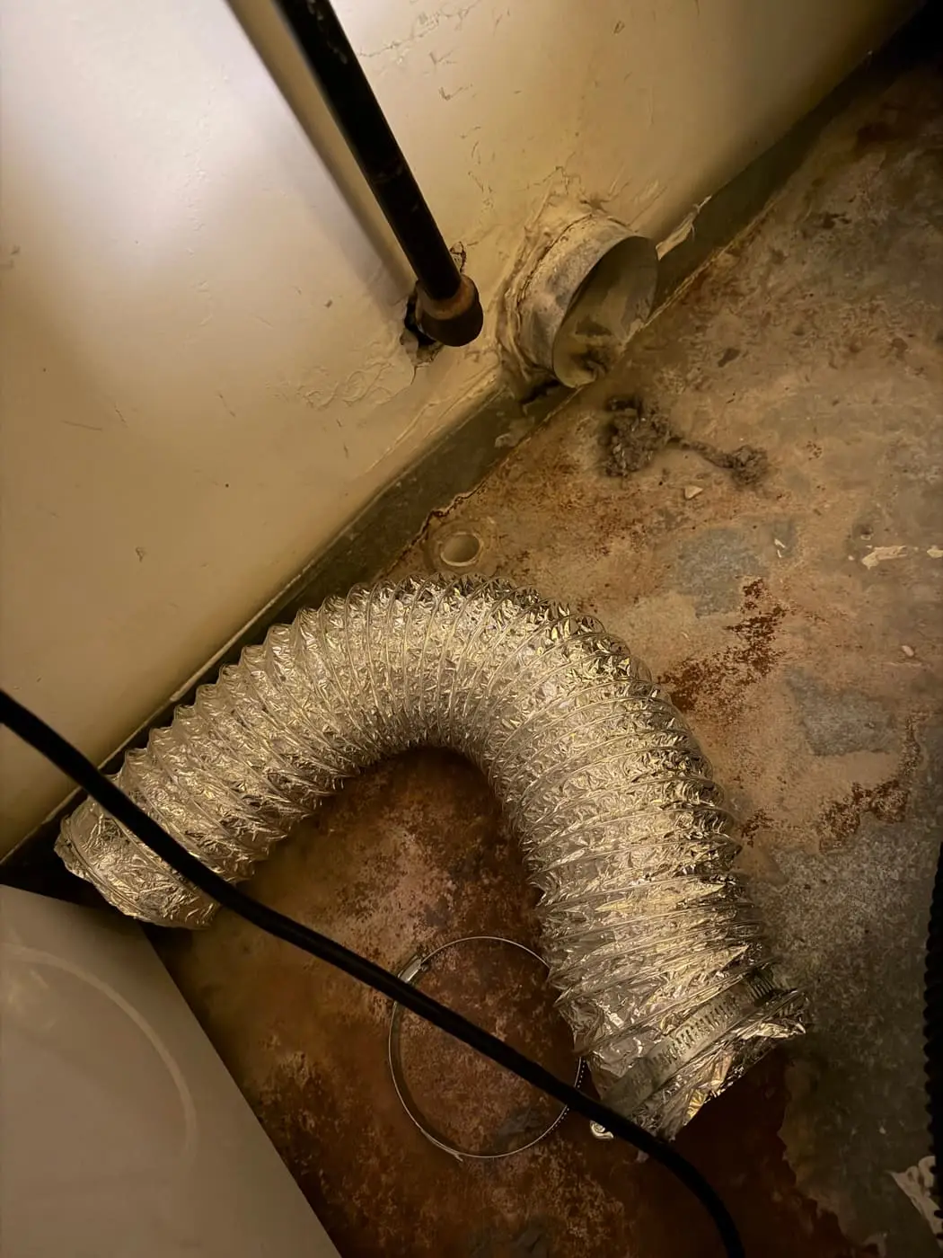 A foil flexible dryer hose lying on the floor, disconnected from the wall vent collar, with lint scattered around it.