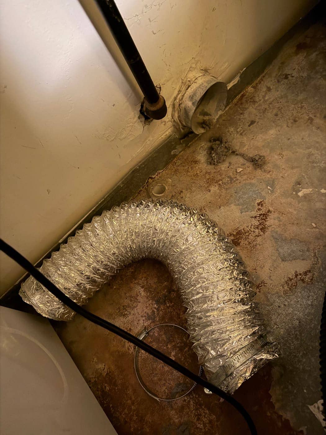 A foil flexible dryer hose lying on the floor, disconnected from the wall vent collar, with lint scattered around it.