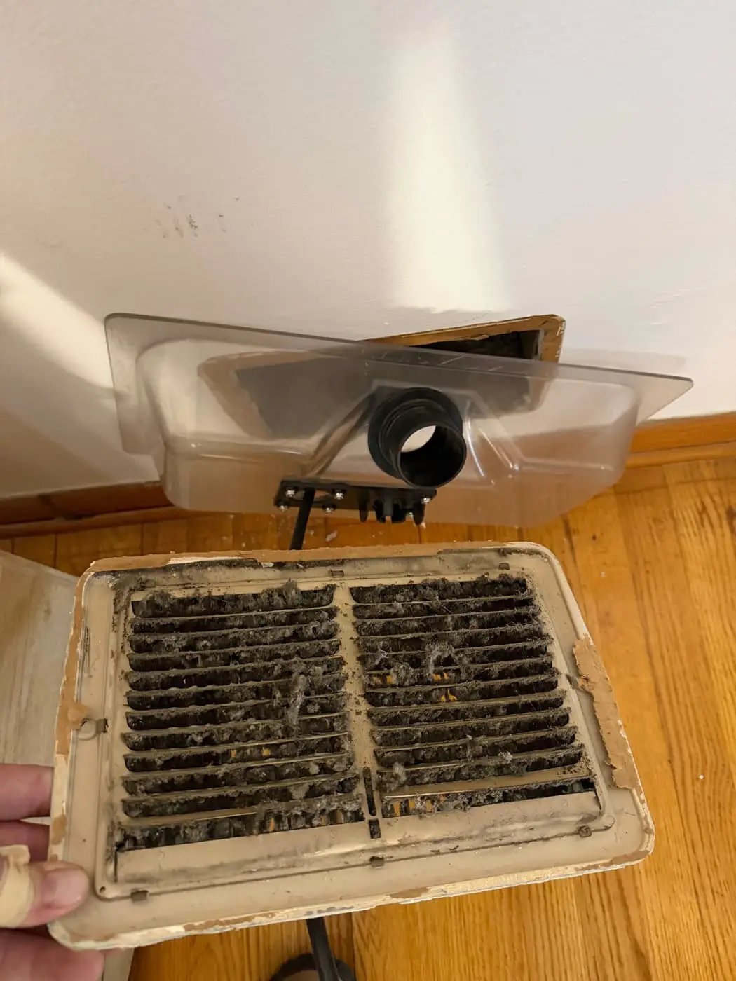 A hand holding a register grille clogged with lint and dust, below a ceiling duct opening fitted with a vacuum hose.
