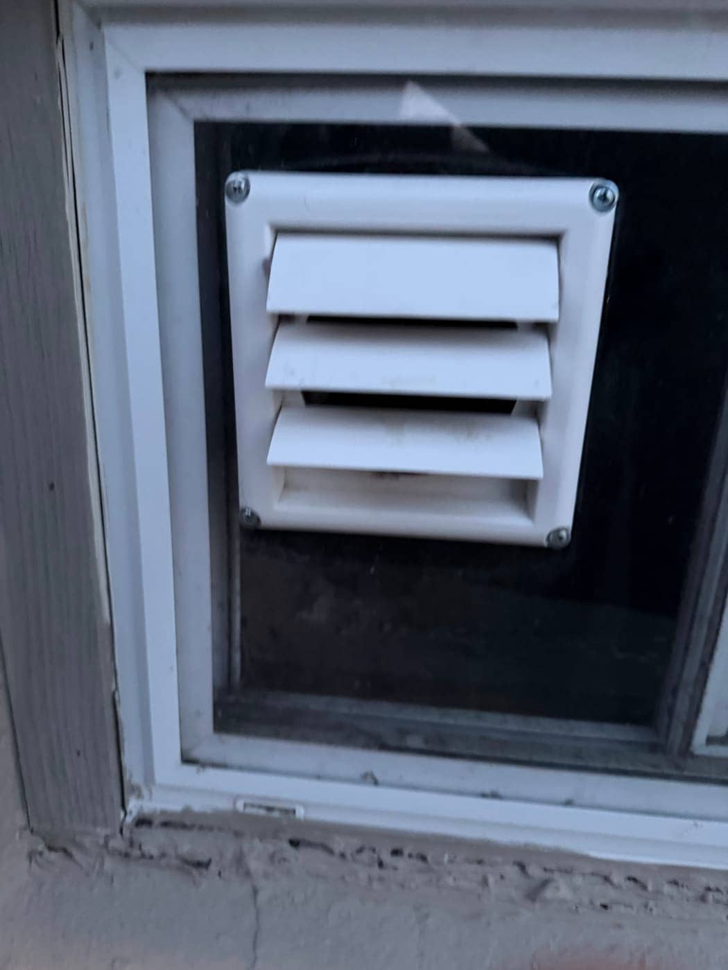 A white louvered vent hood set into a panel that fills a basement window.